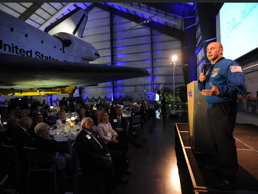 Astronaut Garret Reisman on stage, presenting to gala dinner guests under the space shuttle Endeavour
