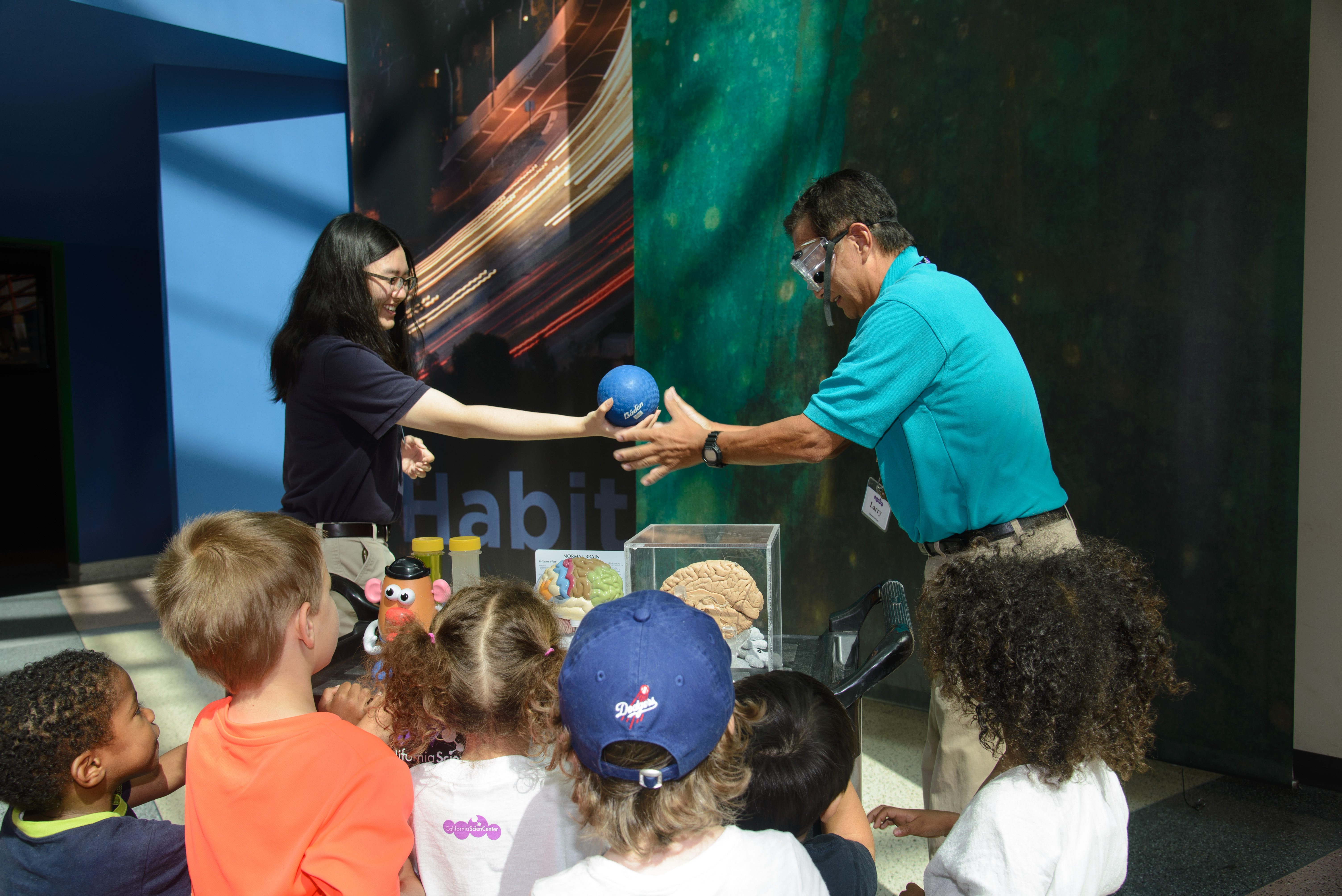 cation team member and Volunteer demonstrate science concept at a cart by passing each other a small blue rubber ball while group of kids watch