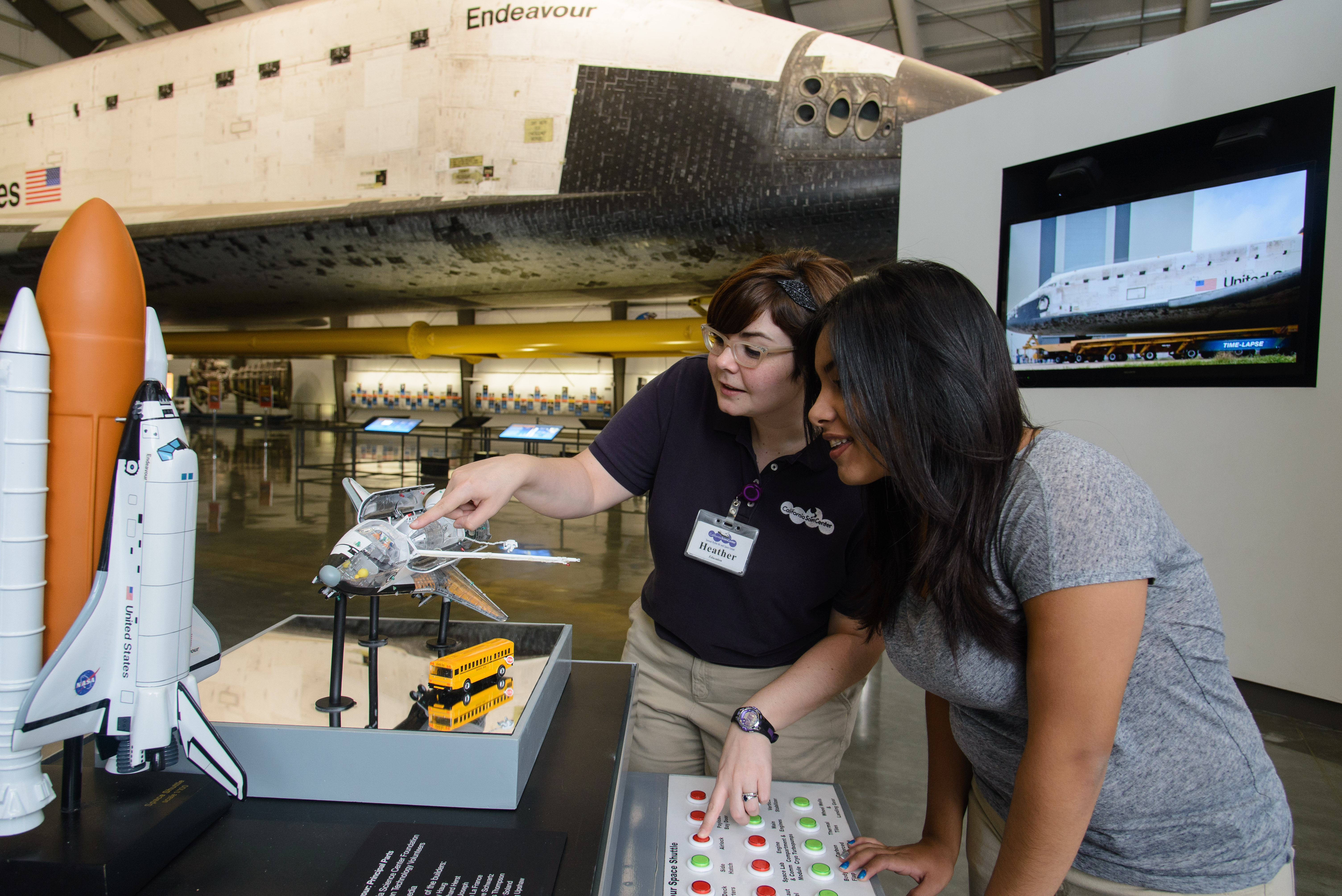 Education staff member points to small-scale model space shuttle with open payload bay as guest observes in the Samuel Oschin Pavilion with space shuttle Endeavour in the background