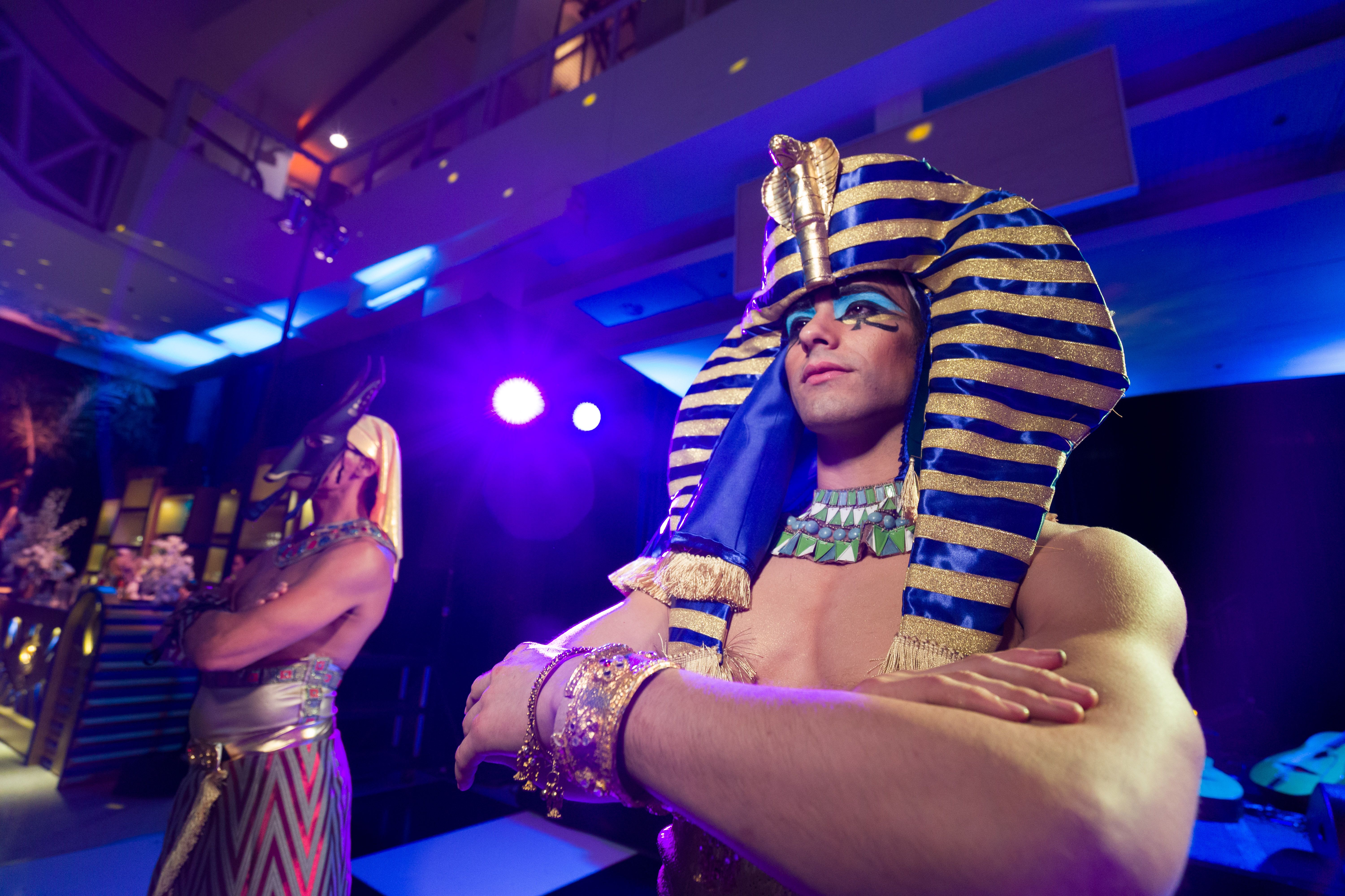 Two male costumed entertainers stand cross armed overlooking Discovery Ball after party. Shirtless entertainers are lit in blue and wearing Egyptian inspired headwear and clothing.