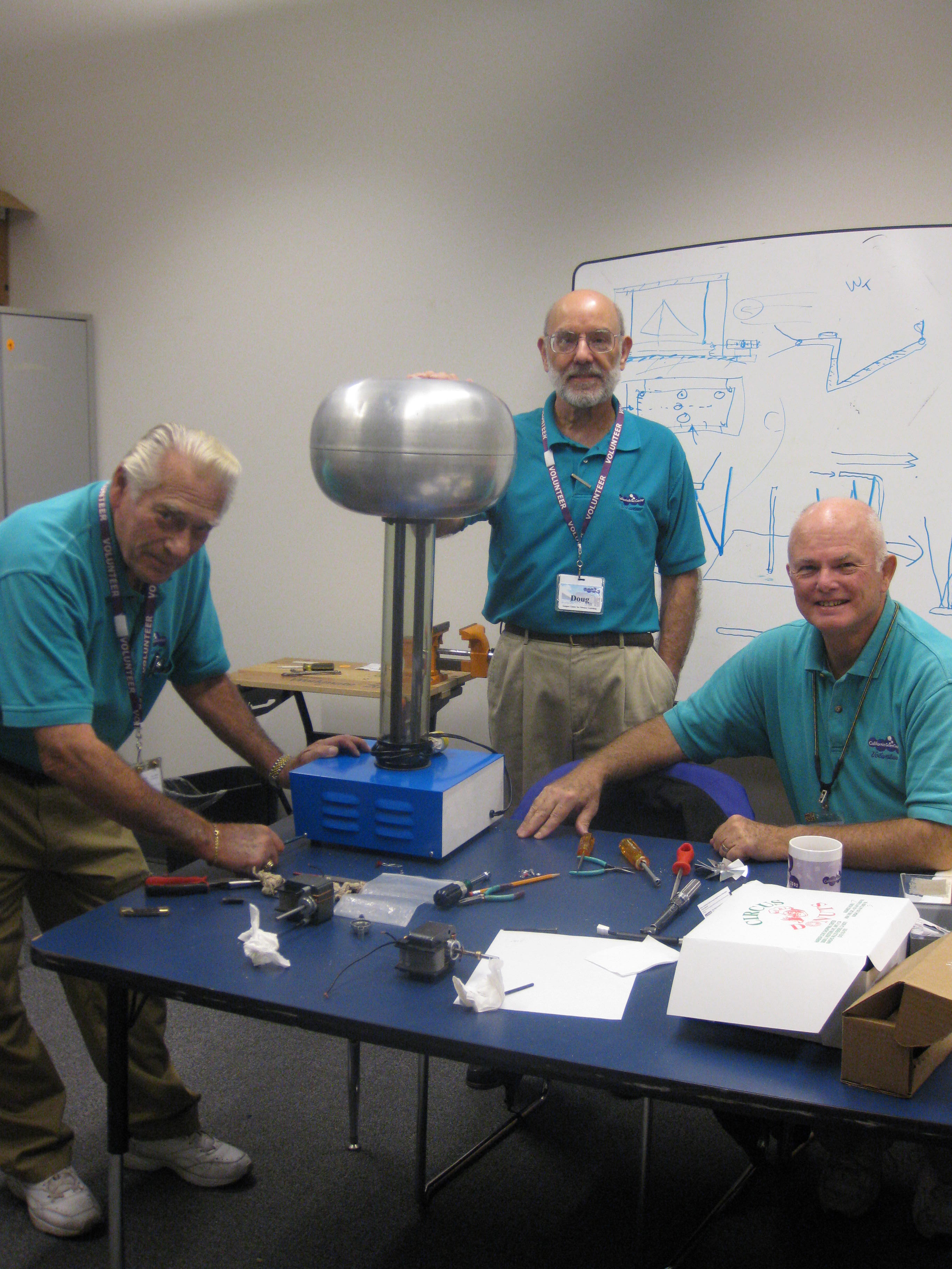 Three Air & Space Volunteers wearing teal polos stand around table with tools and parts while working on Van der Graaf Generator