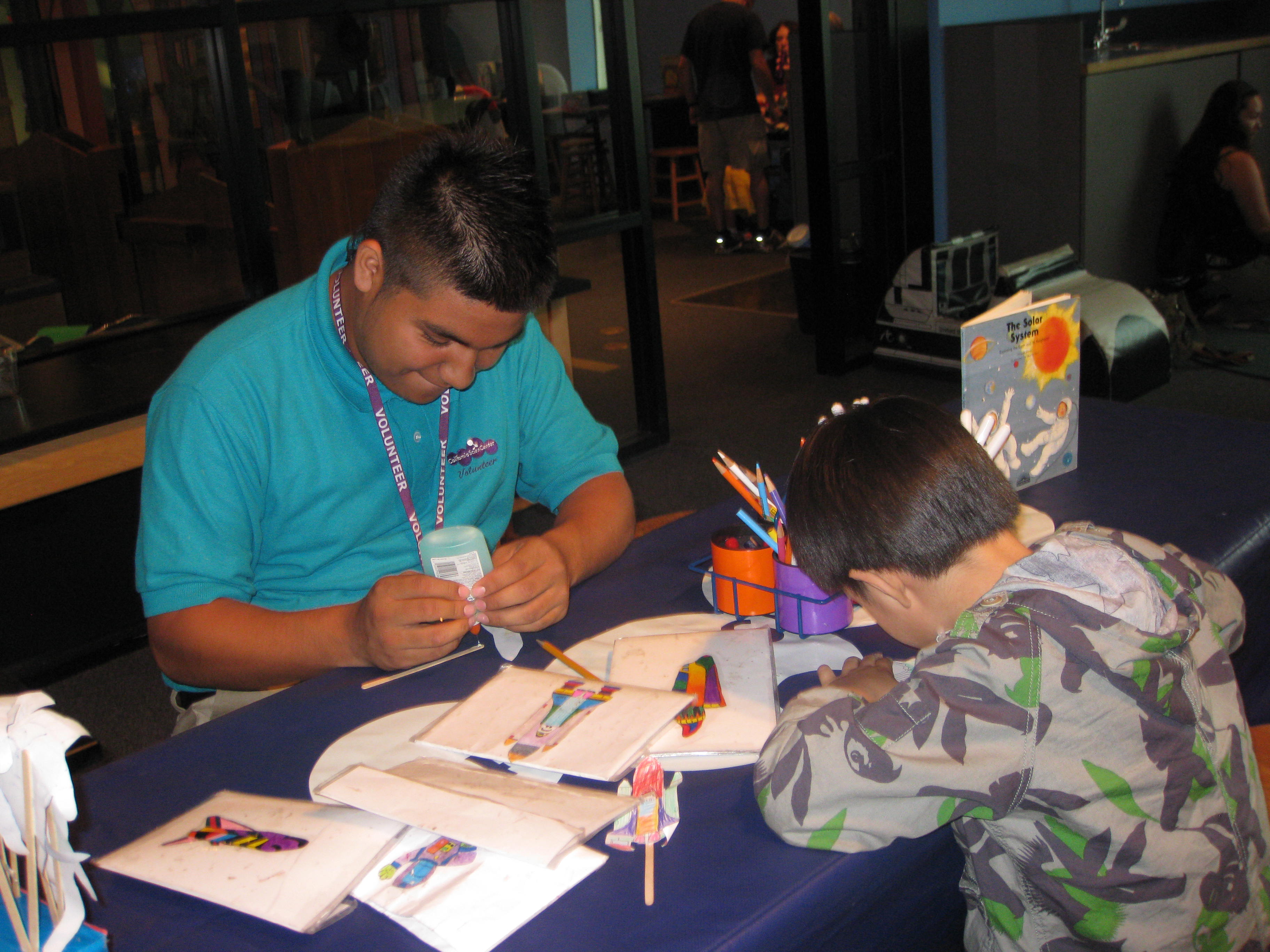 Discovery Room Volunteer wearing teal polo glues Endeavour craft with participating child