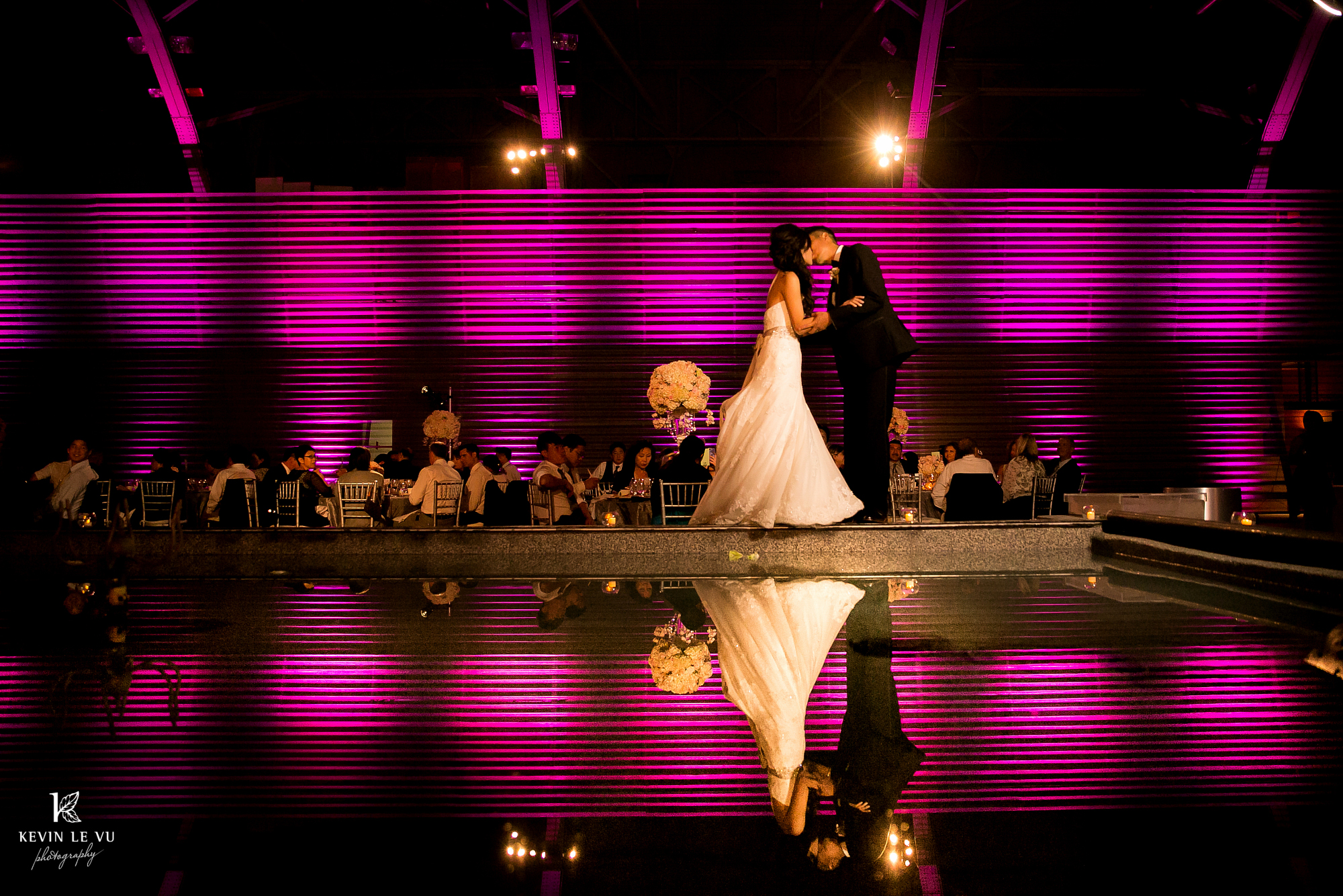 A bride and groom kiss at dusk in front of wedding guests seated at dining rounds and corrugated metal lit in purple. Bride and groom image are reflected in nearby fountain water