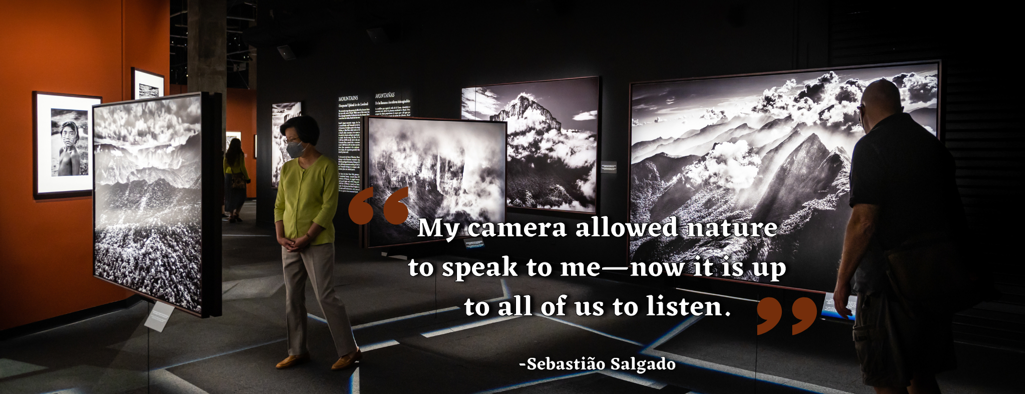 Guests wander through Amazonia exhibit with suspended large-scale photographs. Overlay quote from photographer Sebastiao Salgado reads: My camera allowed nature to speak to me—now it is up to all of us to listen.