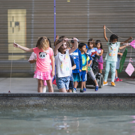 Young kids wait in line to fly their kites near a large fan while at Hands-On Science Camp