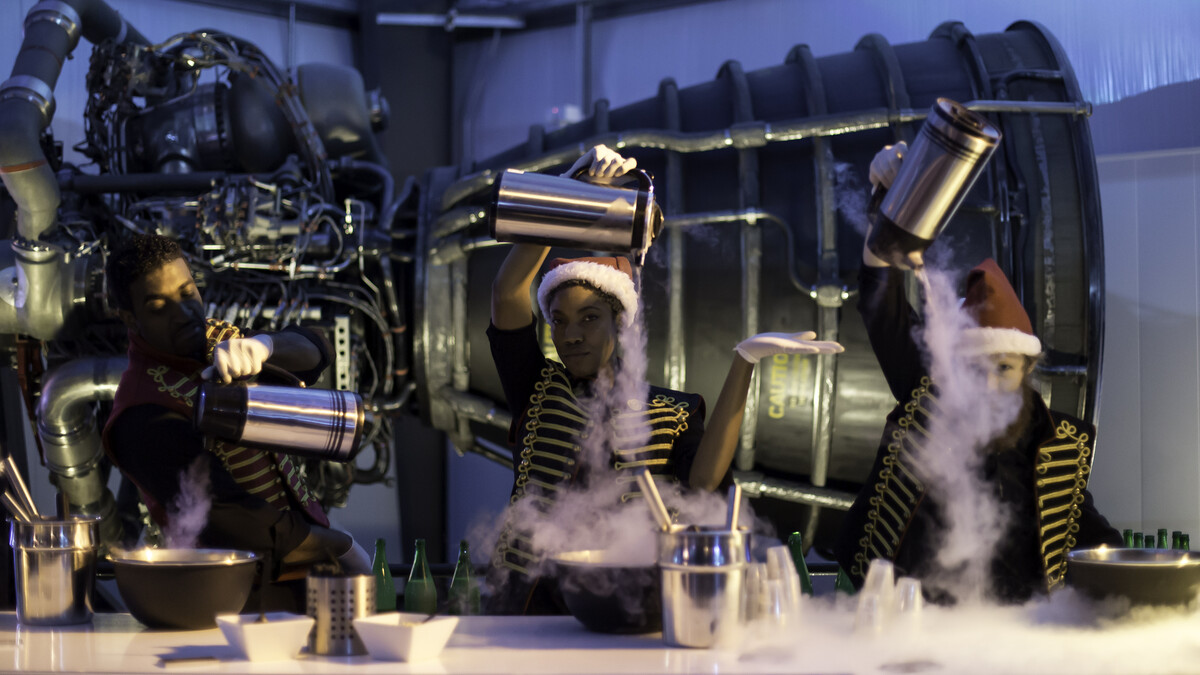 Catering staff in vests and Santa hats pouring liquid nitrogen into ice cream bowls in front of the Space Shuttle Main Engine for a Holiday Party