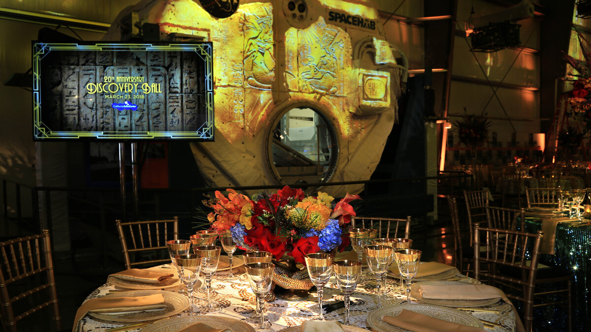 A banquet round with gold adorned linen and dinnerware sits in front of the SpacHab exhibit, which is projection mapped with gold hieroglyphic designs for the King Tut Discovery Ball dinner