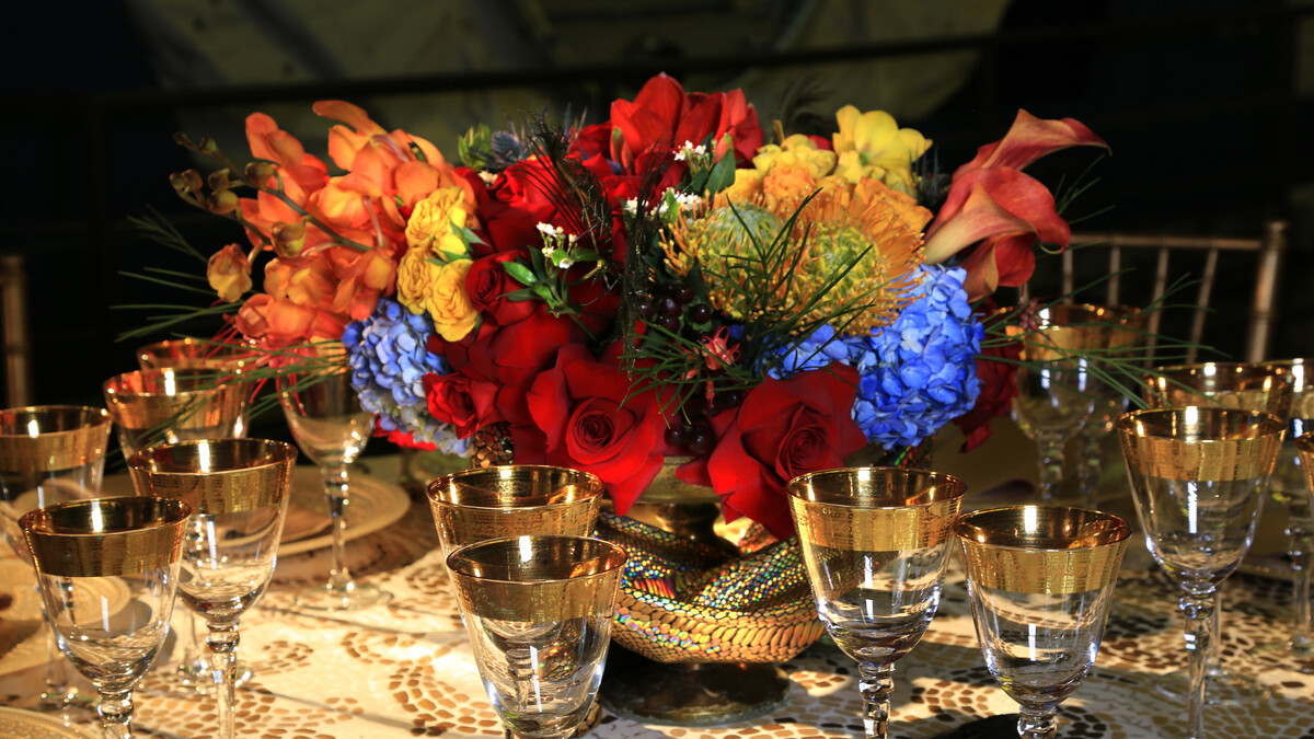 Floral centerpiece with flowers in red, blue, orange, and yellow sits on banquet round dressed for dinner service with gold highlights for the King Tut Discovery Ball