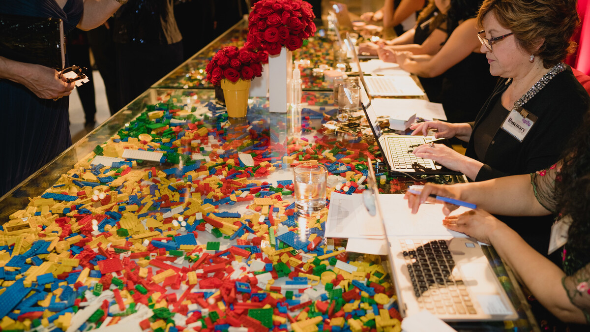 Discovery Ball registration staff working on laptops to check-in guests while sitting at long clear, acrylic table filled with hundreds of LEGO bricks in standard LEGO colors.