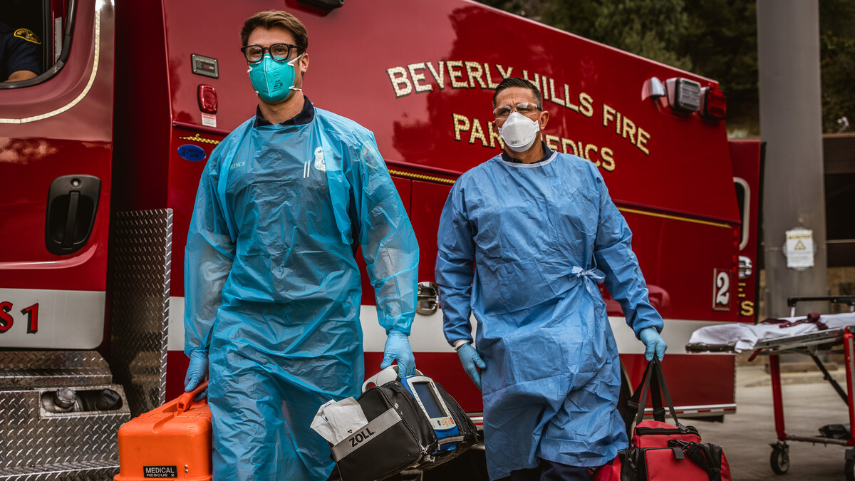 Beverly Hills Fire Department paramedics Jeremy Mack and Austin Prince carry duffel bags of first aid equipment. An ambulance is parked behind them.